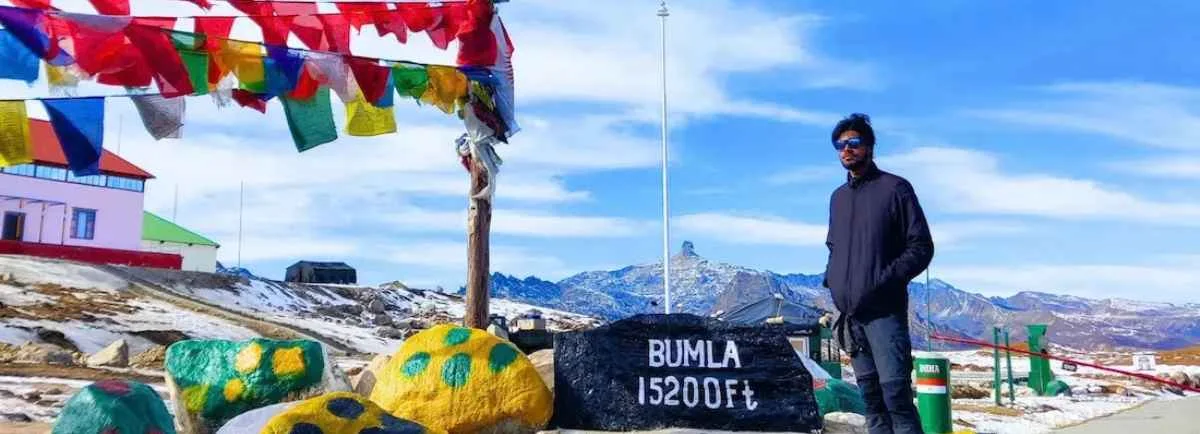 A traveler posing next to a Bum La Pass milestone with prayer flags and snow-capped mountains.