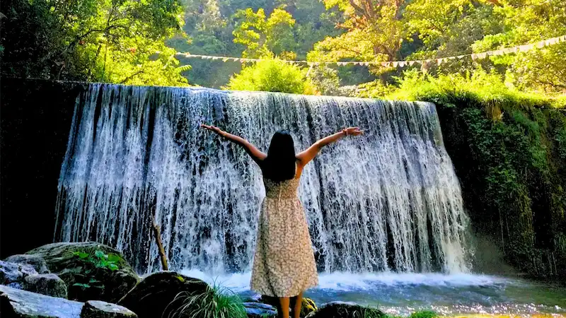 A traveler with arms raised facing Choskorong Waterfall, surrounded by dense green forest.