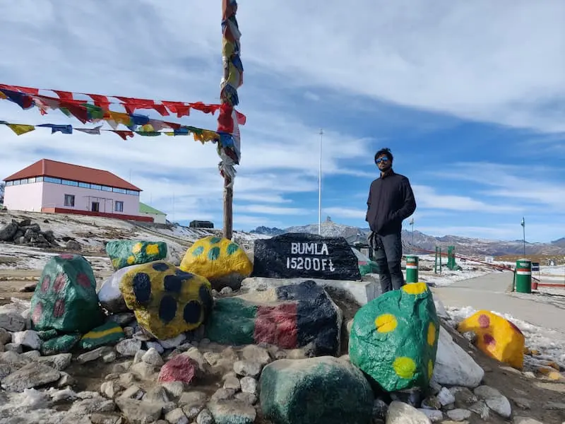 A visitor beside the painted stone marker reading BUMLA 15200 Ft at Bum La Pass near Tawang, prayer flags overhead