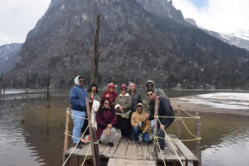 Visitors on a wooden jetty at Sangetsar Lake near Tawang, with bare tree trunks in the water and patchy snow around