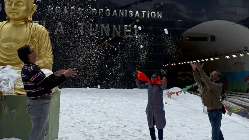 Travelers throwing snow in the air in front of the Sela Tunnel portal near Tawang, with prayer flags and a golden Buddha statue