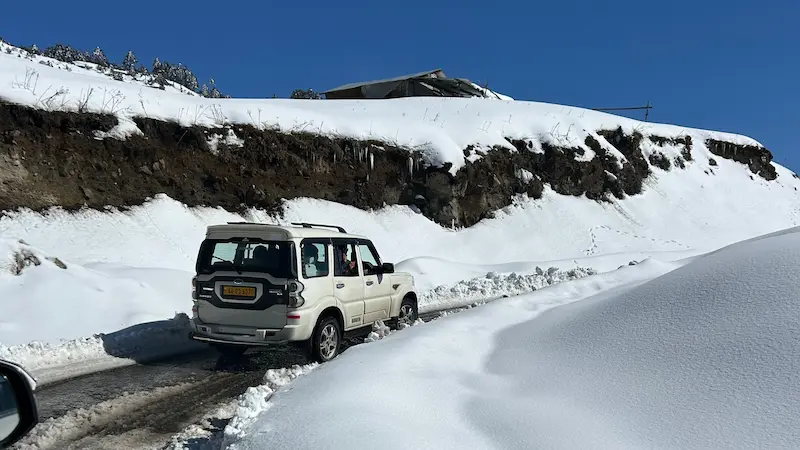 A white SUV on a snow-cleared road cut between deep snowbanks near Tawang under a clear blue sky