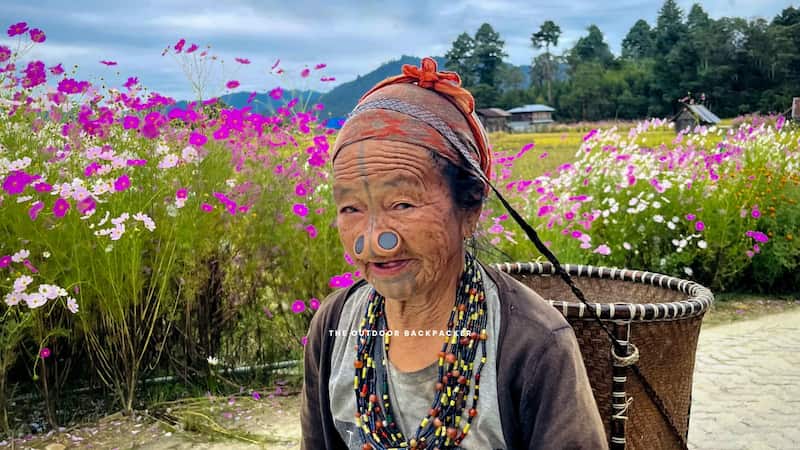 Apatani elder with traditional facial tattoos and nose plugs in a cosmos flower field, Ziro Valley, Arunachal Pradesh