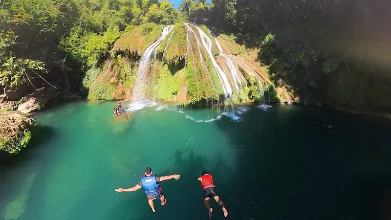 Two travelers jumping into the turquoise pool below Krem Chympe waterfall in Meghalaya.