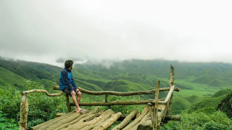 A traveler sitting on a wooden viewpoint deck overlooking the rolling green hills of Dzukou Valley.