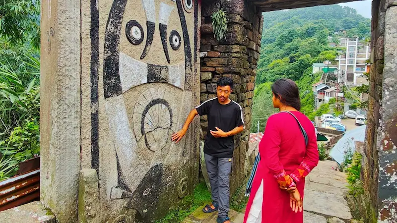 A guide pointing out the carved stone gate at the entrance to Khonoma village to a traveler.