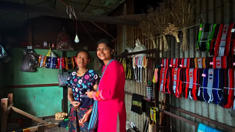 Two women inside a traditional handloom weaving shed in Khonoma, surrounded by colorful woven shawls and textiles.