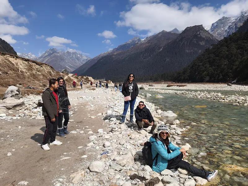 A group of travelers by the clear river in Yumthang Valley, North Sikkim, surrounded by mountains.