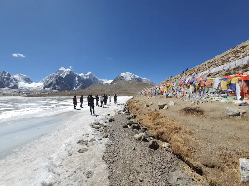 Visitors walking along the frozen edge of Gurudongmar Lake in North Sikkim, prayer flags lining the shore.