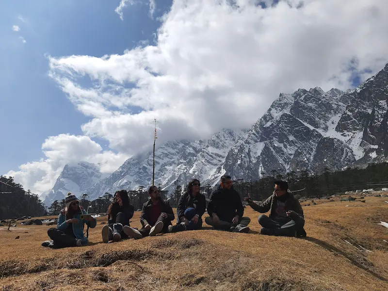 Travelers sitting together on the grass in Yumthang Valley, North Sikkim, with snow-capped peaks and clouds behind them.